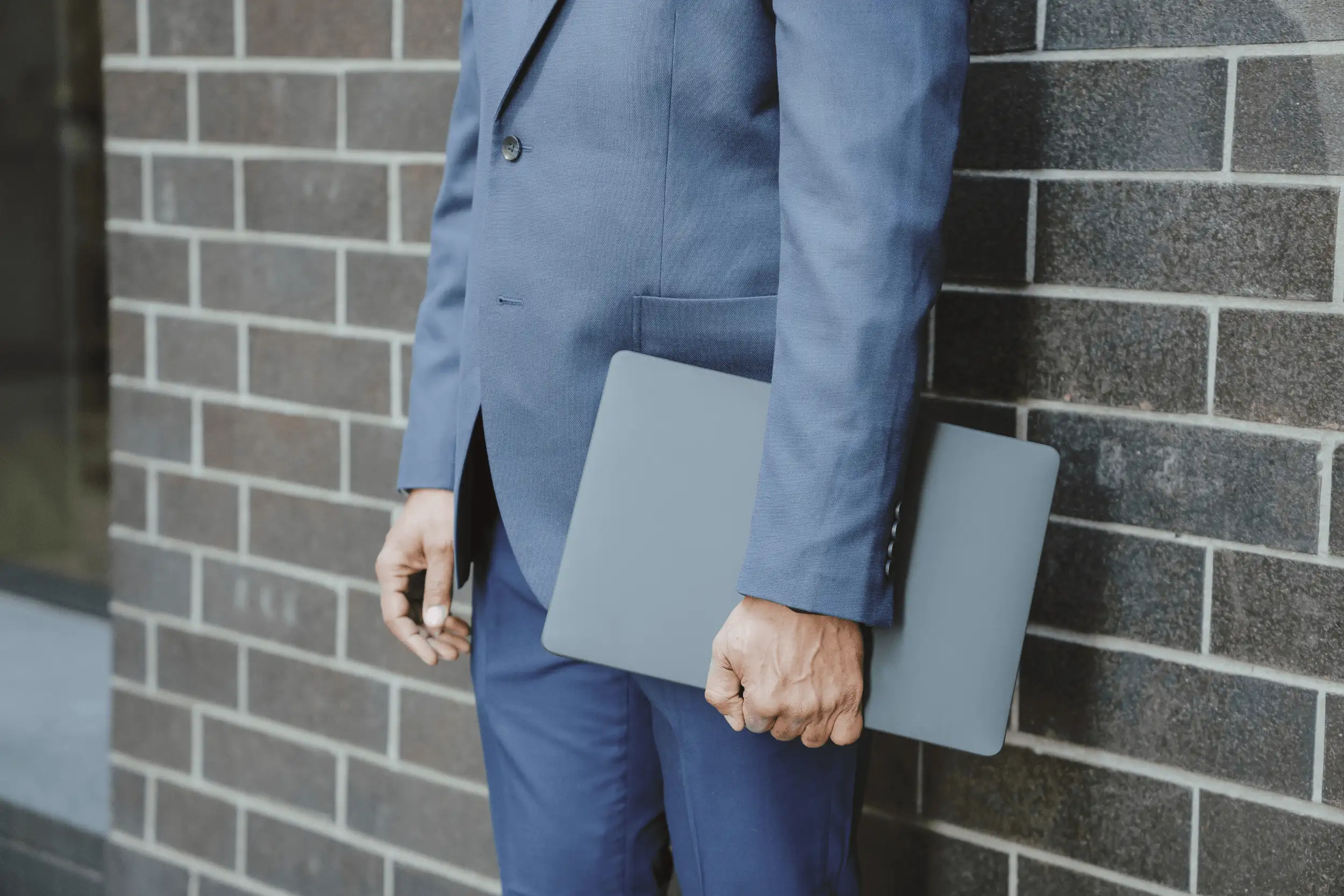 Mann im Anzug steht vor einer Backsteinwand, der ein Laptop in der linken Hand hält.